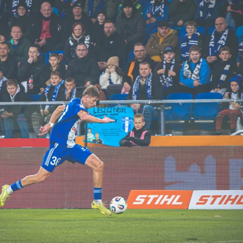 Na zdjęciu młody piłkarz w niebieskim stroju biegnie w kierunku piłki, a w tle widać pełne trybuny kibiców z szalikami.