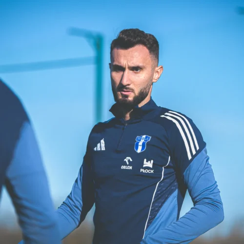 Na zdjęciu widać mężczyznę w sportowej odzieży, który stoi na boisku i wydaje się być skupiony na rozmowie lub treningu.