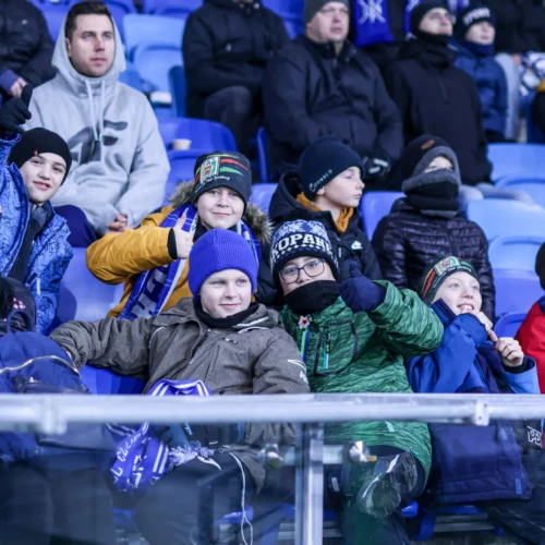 Na zdjęciu widać grupę dzieci i dorosłych w kufrze, którzy z radością kibicują, uśmiechając się i trzymając thumbs up na tle stadionu.