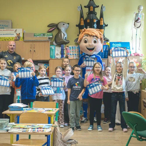 Grupa dzieci stoi w klasie, trzymając kartki, a w tle widoczny jest maskotka oraz nauczyciel.