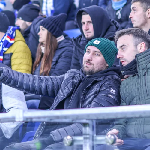 Na zdjęciu widoczni są mężczyźni w ciepłych kurtkach, siedzący na trybunach, z zainteresowaniem rozmawiający i wskazujący w pewnym kierunku.