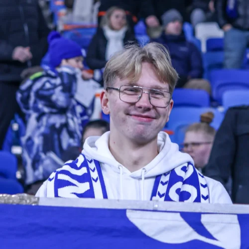 Młody kibic uśmiecha się, trzymając niebieską flagę w tłumie na trybunach stadionu.