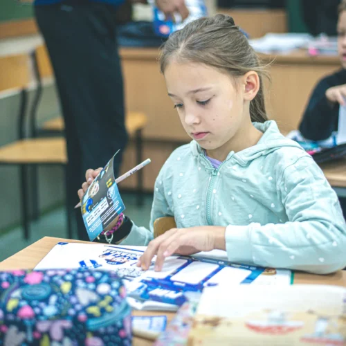 Na zdjęciu widać dziewczynkę w szarej bluzie, która skupiona rysuje lub koloruje w klasie, podczas gdy w tle widać innych uczniów.