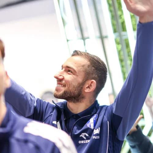 Mężczyzna w niebieskiej odzieży sportowej z uśmiechem macha ręką w górę w jasnym otoczeniu.