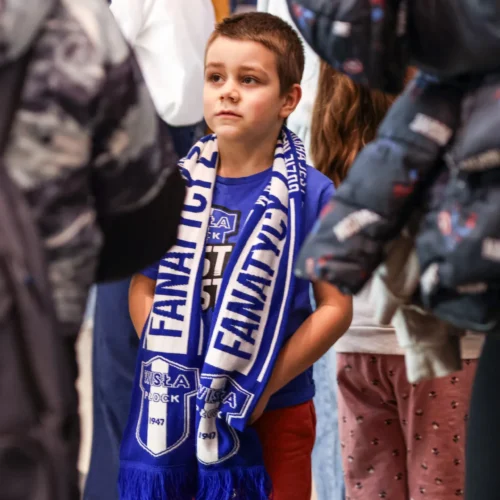 Na zdjęciu widoczny jest mały chłopiec w niebieskiej koszulce oraz szaliku z logo klubu sportowego, który stoi w tłumie i wygląda na zamyślonego.