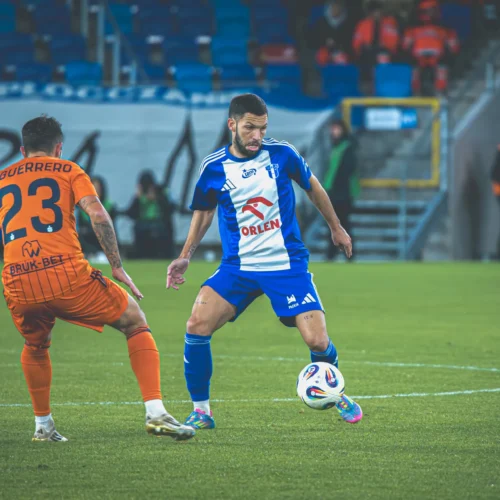 Na zdjęciu dwóch piłkarzy podczas meczu, jeden z nich w niebieskim stroju zmierza w kierunku piłki, a drugi w pomarańczowym stroju próbuje go zablokować.