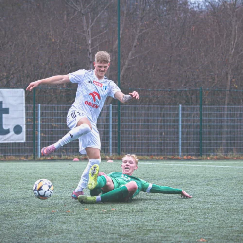 Na zdjęciu widać dwóch młodych piłkarzy w trakcie meczu, gdzie jeden z nich wykonuje zwód, a drugi leży na murawie po próbie interwencji.