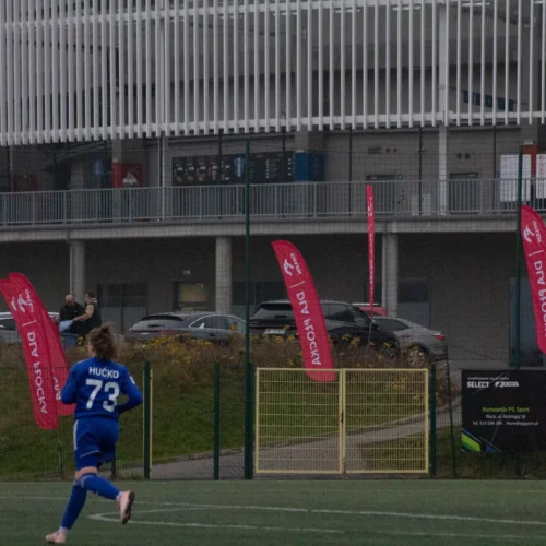 Na zdjęciu widać młodego piłkarza w niebieskim stroju z numerem 73, biegnącego w stronę bramki na boisku sportowym, w tle z flagami reklamowymi i budynkiem.
