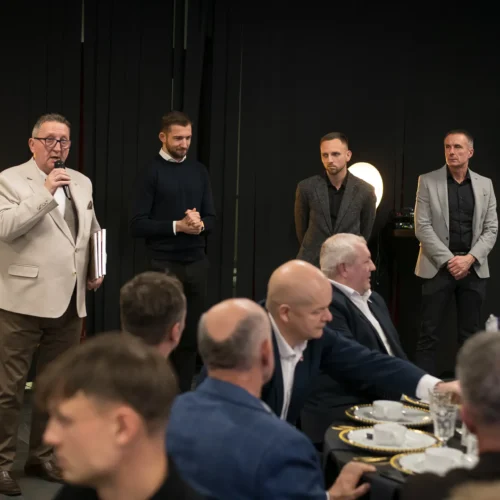 Na zdjęciu widać grupę mężczyzn przy stole, a jeden z nich, w jasnej marynarce, przemawia do zgromadzonych gości.