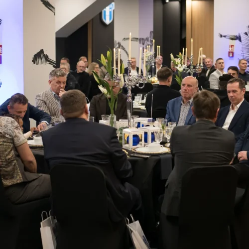 Na zdjęciu widać eleganckie przyjęcie, podczas którego mężczyźni siedzą przy stołach, rozmawiając i spożywając posiłki w dekorowanym wnętrzu.