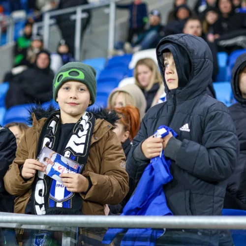 Na zdjęciu widać grupę dzieci i dorosłych na trybunach, wśród których jeden z chłopców trzyma zdjęcie i wygląda na podekscytowanego.