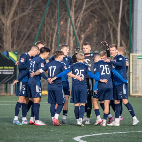Zdjęcie przedstawia grupę piłkarzy w niebieskich strojach, którzy stoją w kręgu i przygotowują się do meczu.