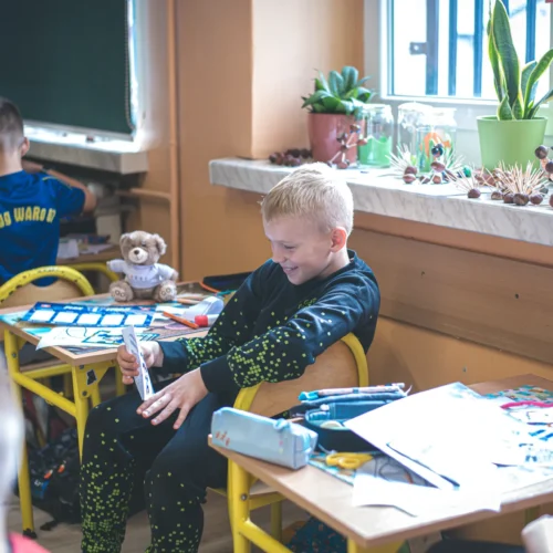 Na zdjęciu widać uśmiechniętego chłopca, który siedzi przy biurku w klasie, trzymając w ręku kartkę papieru, podczas gdy w tle inni uczniowie pracują nad swoimi projektami.