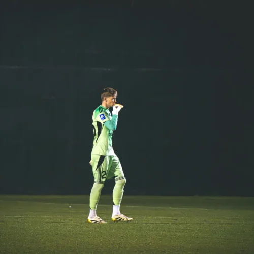 Na zdjęciu widać bramkarza w zielonym stroju, który stoi na boisku piłkarskim, w tle ciemna ściana.