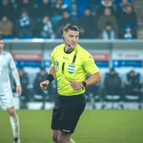 Na zdjęciu widać sędziego piłkarskiego w żółtej koszulce, który kontroluje przebieg meczu na boisku.