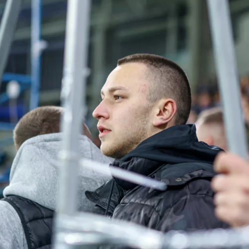 Młody mężczyzna w ciemnej kurtce stojący na trybunach, skupiony na wydarzeniu sportowym.