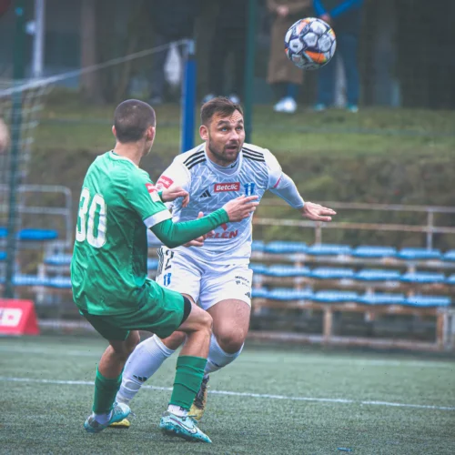 Na zdjęciu widać dwóch piłkarzy podczas meczu, gdzie jeden z nich stara się przejąć piłkę, a drugi unika kontaktu.