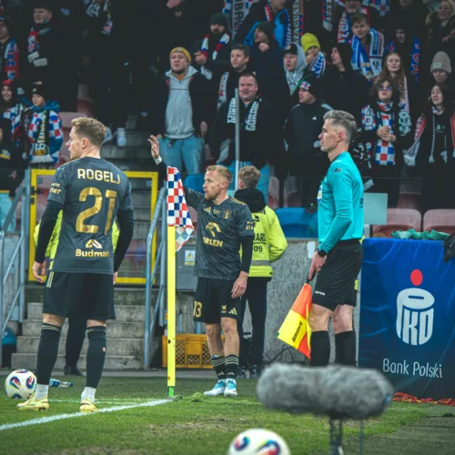 Na zdjęciu widać piłkarzy stojących przy rogu boiska, sędziego oraz kibiców na trybunach, którzy trzymają szaliki drużynowe.