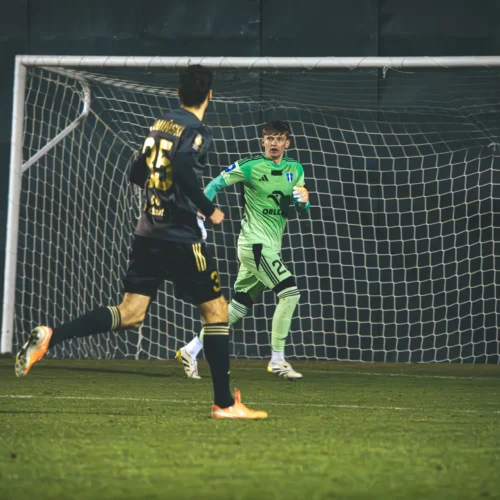 Na zdjęciu widać dwóch piłkarzy na boisku, gdzie bramkarz w zielonym stroju obserwuje nadbiegającego zawodnika w czarnym stroju, a w tle znajduje się bramka piłkarska.