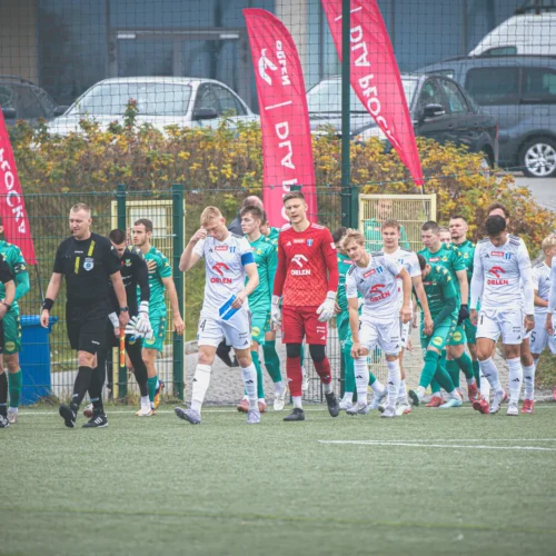 Zawodnicy drużyn piłkarskich i sędziowie wychodzą na boisko, a w tle widoczne są samochody oraz flagi sponsorów.