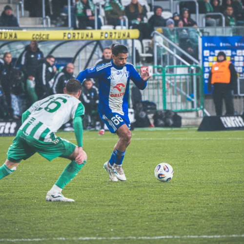 Na zdjęciu widać dwóch piłkarzy w trakcie meczu, z zawodnikiem w niebieskim stroju, który biegnie w kierunku piłki, podczas gdy przeciwnik w zielonym stroju stara się go zatrzymać.