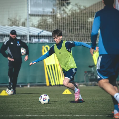 Na zdjęciu widać młodego piłkarza w zielonej koszulce treningowej, który biegnie za piłką na boisku, z trenerem i innymi zawodnikami w tle.