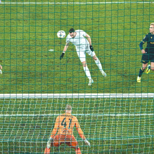 Na zdjęciu znajduje się moment meczu piłkarskiego, gdzie piłka zmierza w kierunku bramkarza, a zawodnicy w strojach drużynowych starają się o nią walczyć w pobliżu bramki.