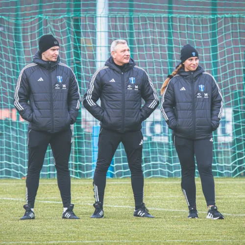 Na zdjęciu widoczna jest trójka osób ubranych w czarne kurtki i spodnie, stojących na boisku piłkarskim przed bramką.