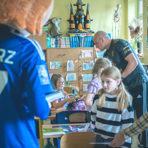 Zdjęcie przedstawia dzieci w klasie, które uczestniczą w aktywności edukacyjnej, z osobą dorosłą w tle oraz maskotką.