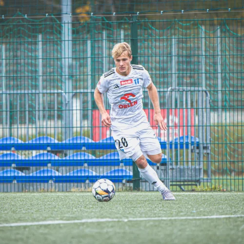 Młody piłkarz w białym stroju, biegnący za piłką na boisku sportowym, w tle z ogrodzeniem i niebieskimi trybunami.