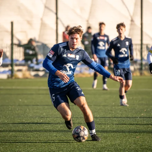 Na zdjęciu widać młodzieżową drużynę piłkarską grającą na boisku, skupioną na akcji przy piłce.