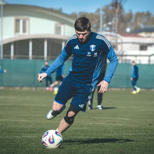 Na zdjęciu widoczny jest piłkarz w niebieskim stroju, który kopie piłkę na boisku treningowym, w tle widać innych zawodników.