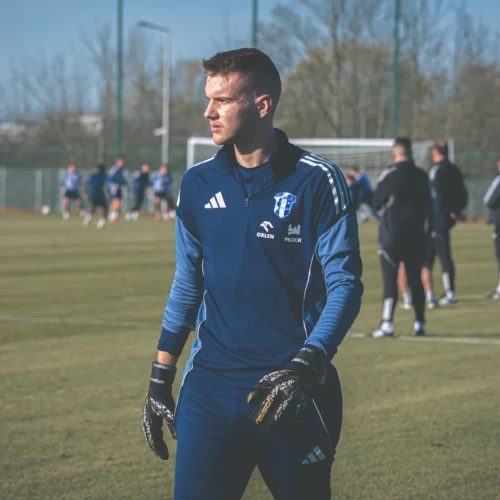 Zawodnik w niebieskiej odzieży sportowej i rękawicach bramkarskich obserwuje trening drużyny na boisku.