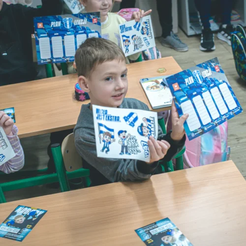 Na zdjęciu widać grupę dzieci w klasie, które trzymają w rękach materiały edukacyjne, w tym plan lekcji i kolorowe naklejki.