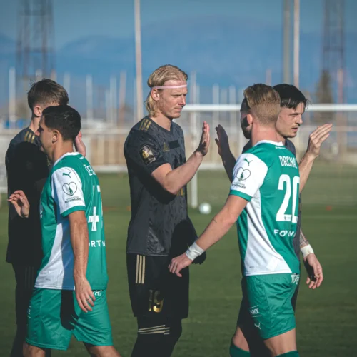 Na zdjęciu widać grupę piłkarzy w różnych strojach, którzy celebrują zakończenie meczu, z gestami uprzejmości i gratulacji.