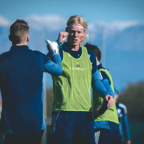 Dwaj piłkarze, jeden w zielonej koszulce treningowej, podnoszą pięść w geście radości w trakcie treningu na tle gór.