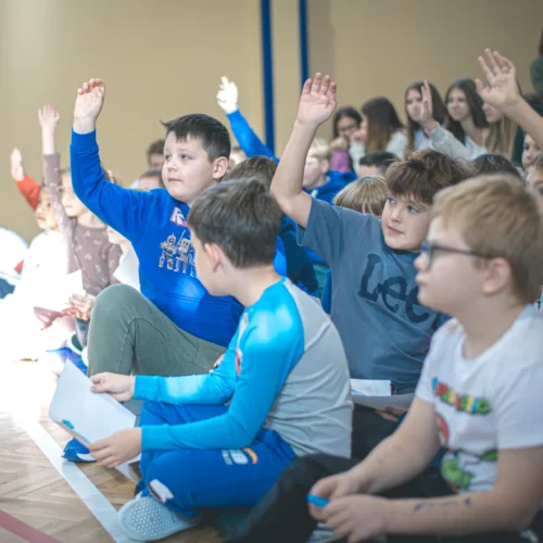 Grupa dzieci na szkolnej sali, większość z uniesionymi rękami, uczestniczy w aktywności lub dyskusji.
