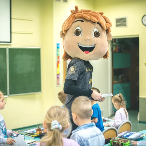 W klasie dziecięcej postać w dużej, uśmiechniętej masce zwierzęcia rozmawia z uczniami siedzącymi przy biurkach.