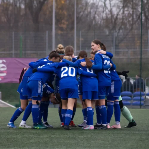 Zawodniczki drużyny piłkarskiej w niebieskich strojach zbierają się w krąg przed rozpoczęciem meczu.