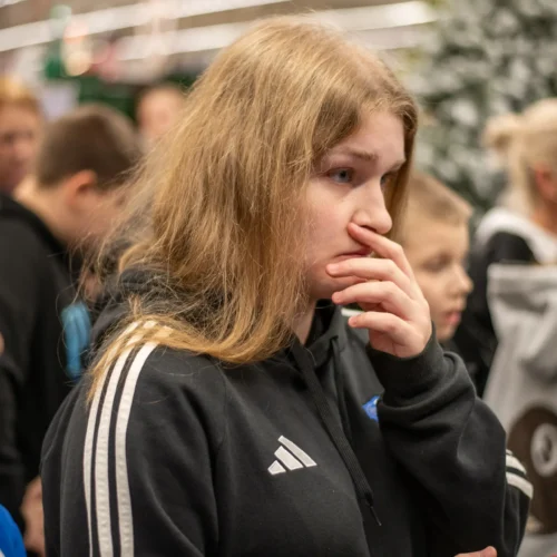 Zdjęcie przedstawia młodą osobę z długimi włosami, która z zamyśloną miną trzyma rękę przy ustach, w otoczeniu innych osób w tle.