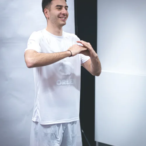 Mężczyzna w białym stroju sportowym, z logo "ORLEN", uśmiecha się i wykonuje gest rąk w studio fotograficznym.