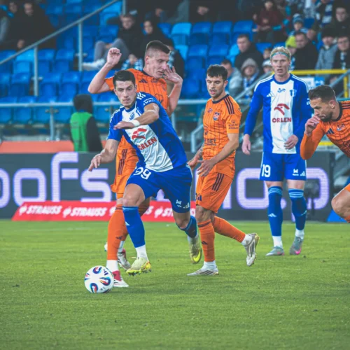 Na zdjęciu widać zawodników drużyn piłkarskich w trakcie meczu, walczących o piłkę na boisku.