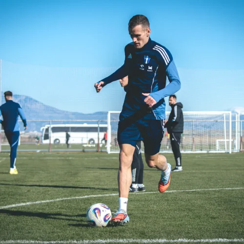 Mężczyzna w sportowym stroju biegnie po boisku piłkarskim, a w tle widać innych zawodników oraz góry.