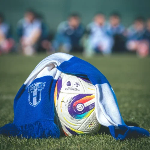 Na zdjęciu znajduje się piłka nożna owinięta niebiesko-białym szalikiem z logo Wisły Płock, na tle zamblurzonej grupy dzieci w strojach sportowych.