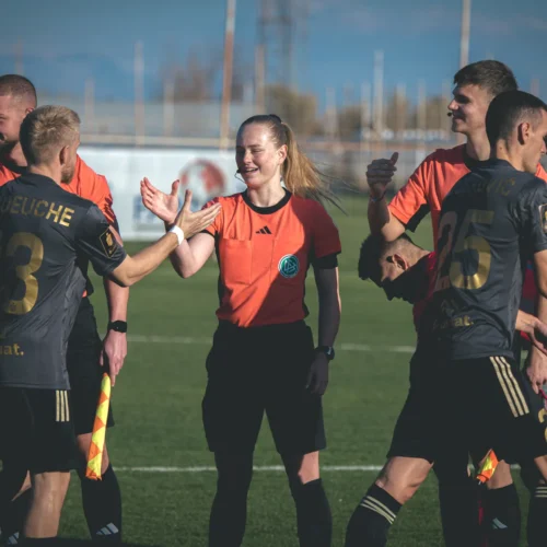 Na zdjęciu znajduje się zawodnik i sędzina na boisku piłkarskim, witający się z innymi graczami w tle.