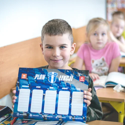 Uśmiechnięty chłopiec trzyma w rękach planszę do planowania lekcji, podczas gdy w tle widać innych uczniów w klasie.