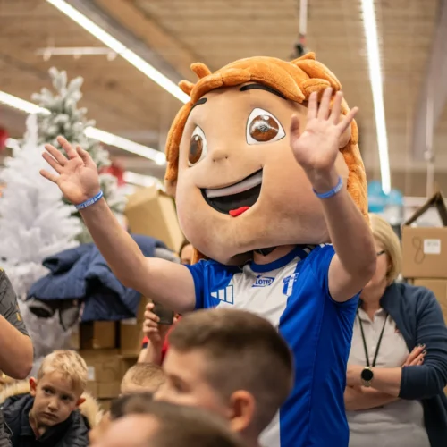 Na zdjęciu widać osobę w kostiumie maskotki z dużą głową, która radośnie macha rękami, a w tle znajdują się dzieci i dekoracje świąteczne.