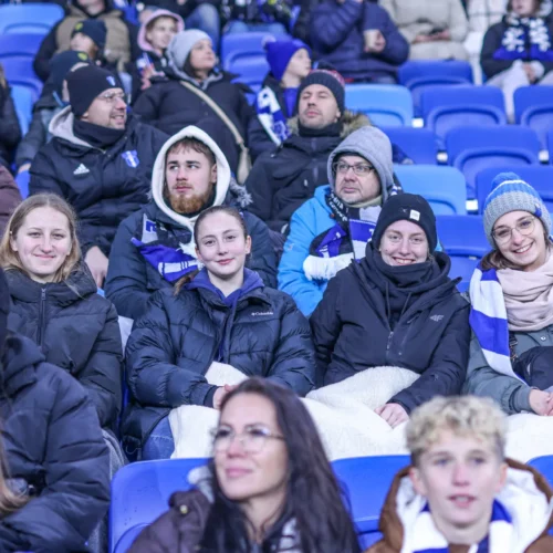 Grupa kibiców w ciepłych kurtkach i szalikach siedzi na trybunach, otoczona przez innych fanów, w atmosferze sportowego wydarzenia.