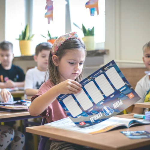 Na zdjęciu znajduje się klasa, w której dzieci siedzą przy biurkach, a dziewczynka z przodu uważnie czyta materiał edukacyjny, podczas gdy inne dzieci zajęte są swoimi zajęciami.