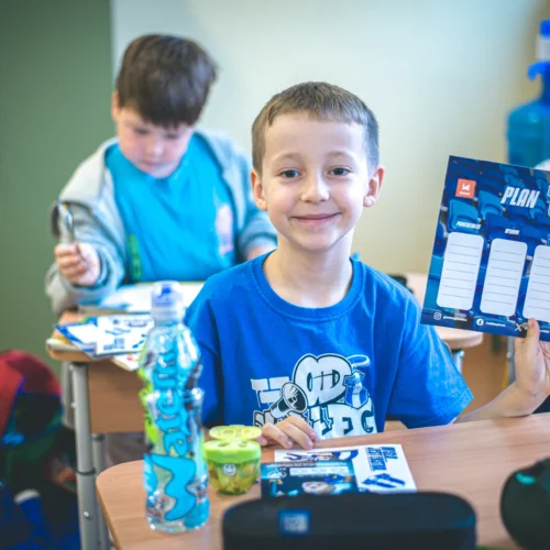 Uśmiechnięty chłopiec w niebieskiej koszulce trzyma w ręku plan lekcji, podczas gdy w tle widać dwóch innych uczniów w szkolnej klasie.